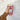 A hand holding a clear plastic cup with a cold coffee drink, wrapped in a maroon and white gingham fabric cup sleeve decorated with cute illustrations of cows, wheat, food, and a teddy bear. The sleeve features a white label that reads "The Love Mich Collection."