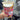 A hand holding a clear plastic cup with a cold drink, wrapped in a maroon and white gingham fabric cup sleeve decorated with cute illustrations of cows, wheat, food, and a teddy bear. The sleeve features a white label that reads "The Love Mich Collection."